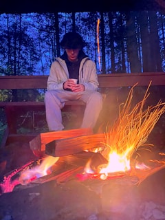 A person is sitting under a wooden shelter in a forest setting, illuminated by the warm glow of a campfire. The individual is wearing casual clothing and appears to be holding a cup. The surrounding trees are silhouetted against a deep blue twilight sky, providing a tranquil and serene backdrop.