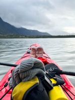 A kayak with various items on top floats on a calm lake. In the foreground, a grey beanie, a yellow dry bag, and a paddle are visible. In the distance, the silhouette of mountains is partially obscured by clouds, and the shore is lined with trees.