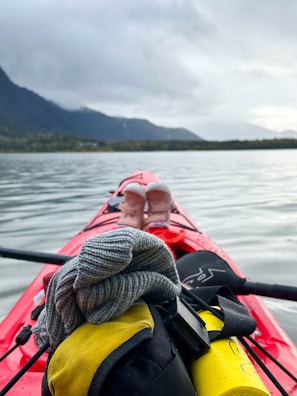 A kayak with various items on top floats on a calm lake. In the foreground, a grey beanie, a yellow dry bag, and a paddle are visible. In the distance, the silhouette of mountains is partially obscured by clouds, and the shore is lined with trees.