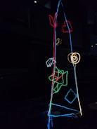 A dark setting featuring a light sculpture composed of colorful neon outlines of various shapes, including a heart, spiral, geometric forms, and abstract patterns. The colors stand out vividly against the black background.