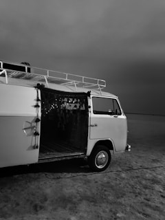 A vintage van is parked on a barren and seemingly endless landscape under a gray sky. The van is partially open, revealing a dimly lit interior with decorative fabric hanging from the door. The scene conveys a sense of solitude and adventure.