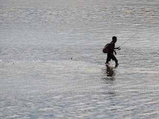 A person is walking through shallow water, holding a fishing rod. The individual appears to be wearing a backpack and is silhouetted against the reflective water surface.