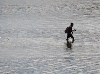 A person is walking through shallow water, holding a fishing rod. The individual appears to be wearing a backpack and is silhouetted against the reflective water surface.