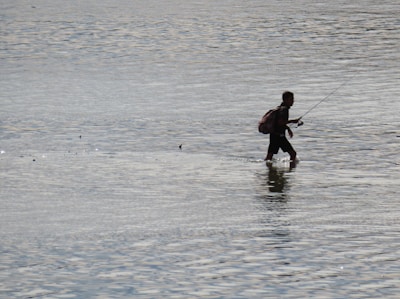 A person is walking through shallow water, holding a fishing rod. The individual appears to be wearing a backpack and is silhouetted against the reflective water surface.