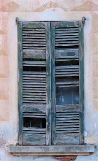 A set of weathered and aged wooden shutters cover a window, showing signs of wear with peeling paint and broken slats. The wood has a mix of green and natural wood tones, and the window behind features lace curtains, adding a touch of delicacy to the rustic scene. The surrounding wall shows signs of aging with its textured, faded plaster.