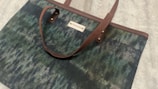A bag with a patterned design featuring shades of green and blue, accented with brown leather straps and a small label on the front.