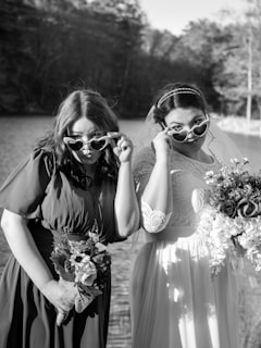 Two women are standing outdoors near a body of water, holding bouquets and wearing heart-shaped sunglasses. One is in a bridal gown with lace details, while the other is in a darker bridesmaid dress. Both are making playful expressions and posing with their sunglasses.