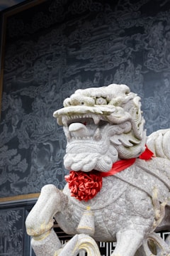 A stone statue of a lion adorned with a red silk ribbon around its neck stands prominently. The intricate details of the statue capture the lion's mane and facial expressions. In the background, there is a detailed mural depicting clouds and mythological figures, adding a rich historical ambiance.