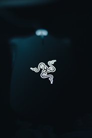 A luminous, stylized triple-headed snake logo is set against a deep, dark background. The logo is bright and eye-catching, emitting a soft glow that contrasts with the surrounding darkness.