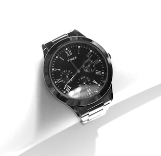 A stylish wristwatch with a metallic band and a black round face featuring Roman numerals and multiple dials, set against a minimalist white background.