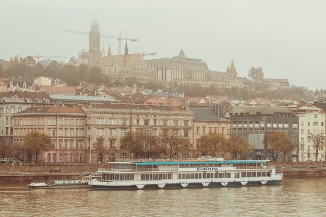 A river cruise ship is anchored along the waterfront, with historic buildings and a church set on a hill in the background. The scene is enveloped in a light mist, adding a soft, ethereal quality.