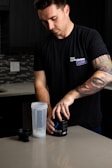 A man with a tattooed arm is standing at a kitchen counter. He is wearing a black t-shirt with text on it and is opening a black container labeled 'BUCKED UP'. A clear shaker bottle with a metal blender ball is placed on the counter beside him.