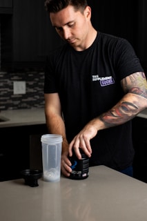 A man with a tattooed arm is standing at a kitchen counter. He is wearing a black t-shirt with text on it and is opening a black container labeled 'BUCKED UP'. A clear shaker bottle with a metal blender ball is placed on the counter beside him.