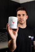 A person is holding up a white container with green and red branding that features a design of deer antlers and a head. The person is standing in front of a dark background and is wearing a black shirt.