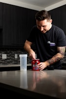 A person with a tattoo on their arm is standing at a kitchen counter, scooping powder from a red container labeled 'NO XPLODE' into a shaker bottle. The person is wearing a dark t-shirt, and the kitchen has dark cabinets and a tile backsplash.