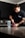 A person with a tattoo on their arm is standing at a kitchen counter, scooping powder from a red container labeled 'NO XPLODE' into a shaker bottle. The person is wearing a dark t-shirt, and the kitchen has dark cabinets and a tile backsplash.