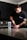 A person with a tattoo on their arm is standing at a kitchen counter, scooping powder from a red container labeled 'NO XPLODE' into a shaker bottle. The person is wearing a dark t-shirt, and the kitchen has dark cabinets and a tile backsplash.