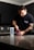 A person with a tattoo on their arm is standing at a kitchen counter, scooping powder from a red container labeled 'NO XPLODE' into a shaker bottle. The person is wearing a dark t-shirt, and the kitchen has dark cabinets and a tile backsplash.