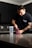 A person with a tattoo on their arm is standing at a kitchen counter, scooping powder from a red container labeled 'NO XPLODE' into a shaker bottle. The person is wearing a dark t-shirt, and the kitchen has dark cabinets and a tile backsplash.