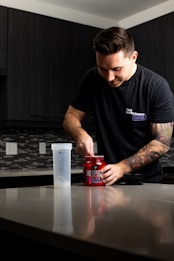 A person with a tattoo on their arm is standing at a kitchen counter, scooping powder from a red container labeled 'NO XPLODE' into a shaker bottle. The person is wearing a dark t-shirt, and the kitchen has dark cabinets and a tile backsplash.