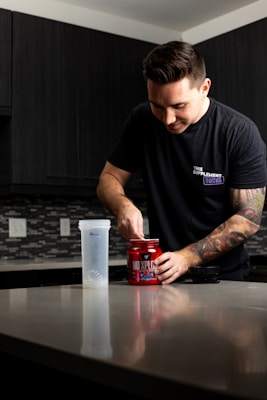 A person with a tattoo on their arm is standing at a kitchen counter, scooping powder from a red container labeled 'NO XPLODE' into a shaker bottle. The person is wearing a dark t-shirt, and the kitchen has dark cabinets and a tile backsplash.