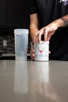 A person is opening a container labeled 'BUCKED UP' on a kitchen counter. Next to it is a transparent shaker bottle. The scene suggests a focus on fitness or supplementation.