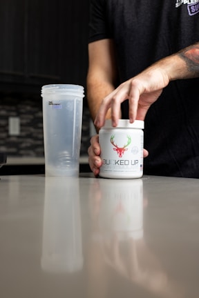 A person is opening a container labeled 'BUCKED UP' on a kitchen counter. Next to it is a transparent shaker bottle. The scene suggests a focus on fitness or supplementation.