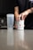 A person is opening a container labeled 'BUCKED UP' on a kitchen counter. Next to it is a transparent shaker bottle. The scene suggests a focus on fitness or supplementation.