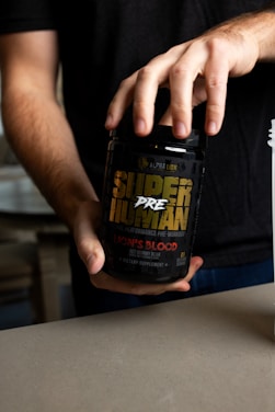 A person holding a black container labeled 'Super Human Pre-Workout' by Alpha Lion. The label has yellow and red text, indicating that it is a dietary supplement with a 'Lion's Blood Red Gummy Bear' flavor. The person's hands are visible, and they are wearing a dark shirt.
