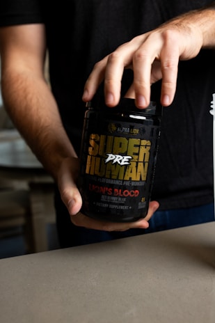 A person holding a black container labeled 'Super Human Pre-Workout' by Alpha Lion. The label has yellow and red text, indicating that it is a dietary supplement with a 'Lion's Blood Red Gummy Bear' flavor. The person's hands are visible, and they are wearing a dark shirt.