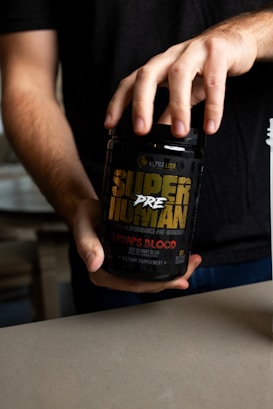 A person holding a black container labeled 'Super Human Pre-Workout' by Alpha Lion. The label has yellow and red text, indicating that it is a dietary supplement with a 'Lion's Blood Red Gummy Bear' flavor. The person's hands are visible, and they are wearing a dark shirt.