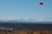 A kiteboarder is gliding over the water, holding onto a bright red kite. The scene is set against a backdrop of a vast body of water with gentle waves. The horizon features mountainous terrain under a clear blue sky with some fluffy, white clouds. In the foreground, dry grass can be seen at the edge of the shore.
