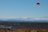 A kiteboarder is gliding over the water, holding onto a bright red kite. The scene is set against a backdrop of a vast body of water with gentle waves. The horizon features mountainous terrain under a clear blue sky with some fluffy, white clouds. In the foreground, dry grass can be seen at the edge of the shore.