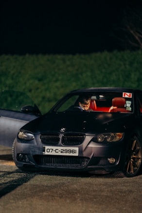 A black BMW car with its door open, parked outdoors at night. The interior is illuminated, showing red seats and a person seated inside. The car has a license plate with the number 07-C-29961 and displays an 'R' plate on the windshield. The background is dark with a hedge visible.