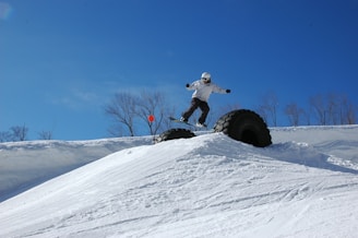 A snowboarder is performing a jump over a large, partially buried rubber tire in a snowy landscape. The sky is clear and blue, and leafless trees stand in the background. The snowboarder is dressed in winter gear, including a helmet and goggles, and appears to be in motion.