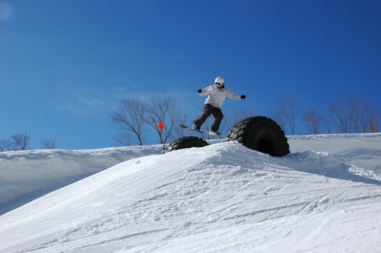 A snowboarder is performing a jump over a large, partially buried rubber tire in a snowy landscape. The sky is clear and blue, and leafless trees stand in the background. The snowboarder is dressed in winter gear, including a helmet and goggles, and appears to be in motion.