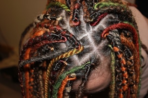 Several braids with vibrant colors, including orange, red, green, and black, tightly woven into the hair. The braids create intricate patterns against the scalp, with each section clearly defined.