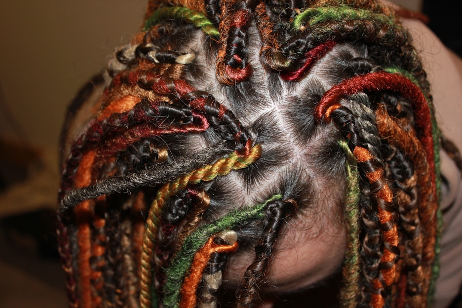 Several braids with vibrant colors, including orange, red, green, and black, tightly woven into the hair. The braids create intricate patterns against the scalp, with each section clearly defined.