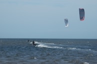 Two kite surfers are gliding over the ocean waves, each attached to colorful kites high in the sky. The sea appears choppy, and the sky is mostly clear with some clouds.