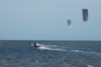 Two kite surfers are gliding over the ocean waves, each attached to colorful kites high in the sky. The sea appears choppy, and the sky is mostly clear with some clouds.