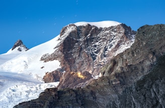 A rugged mountain range with snow-covered peaks under a clear blue sky. The central peak has rough, textured rock surfaces with patches of snow. Visible is a small illuminated cabin or structure nestled among the rocks, emitting a warm, glowing light.