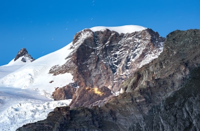 A rugged mountain range with snow-covered peaks under a clear blue sky. The central peak has rough, textured rock surfaces with patches of snow. Visible is a small illuminated cabin or structure nestled among the rocks, emitting a warm, glowing light.