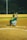 A baseball player in a green jersey squats in a catcher's position on a dirt baseball field at night. Another player is visible in the background, preparing to pitch the ball. The field is illuminated by artificial lights and is surrounded by a fence.