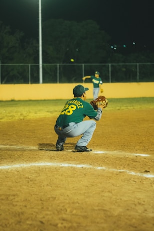 A baseball player in a green jersey squats in a catcher's position on a dirt baseball field at night. Another player is visible in the background, preparing to pitch the ball. The field is illuminated by artificial lights and is surrounded by a fence.