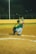 A baseball player in a green jersey squats in a catcher's position on a dirt baseball field at night. Another player is visible in the background, preparing to pitch the ball. The field is illuminated by artificial lights and is surrounded by a fence.