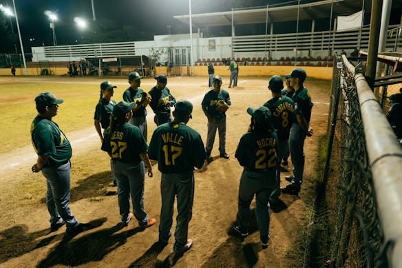 A group of baseball players in green and yellow uniforms gather in a semi-circle on the field, some wearing caps and others holding gloves. The scene takes place in an outdoor stadium with bright lights, a yellow barrier, and protective fencing nearby. The field shows signs of use with patches of dirt and grass.
