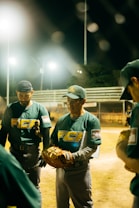 A group of baseball players in green uniforms with text on them gather together at night on a sports field. One player is holding a baseball glove and appears to be speaking or giving instructions. The scene is illuminated by bright stadium lights in the background.