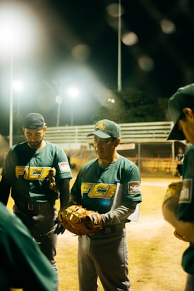 A group of baseball players in green uniforms with text on them gather together at night on a sports field. One player is holding a baseball glove and appears to be speaking or giving instructions. The scene is illuminated by bright stadium lights in the background.