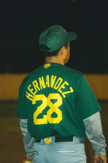 A person wearing a green baseball jersey with the number 28 and the name 'HERNANDEZ' on the back. The individual also wears a matching green baseball cap and grey pants with layered grey sleeves.