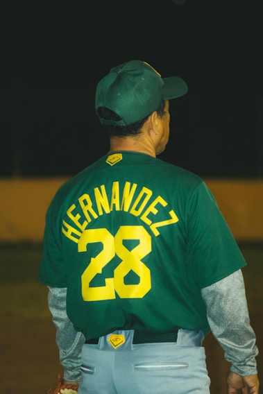 A person wearing a green baseball jersey with the number 28 and the name 'HERNANDEZ' on the back. The individual also wears a matching green baseball cap and grey pants with layered grey sleeves.